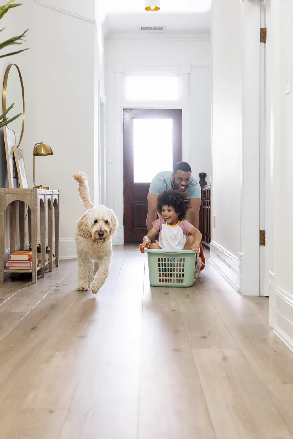 A father pushes his young child in a laundry basket down a hallway while their golden dog trots alongside &mdash; on wide-plank luxury vinyl flooring